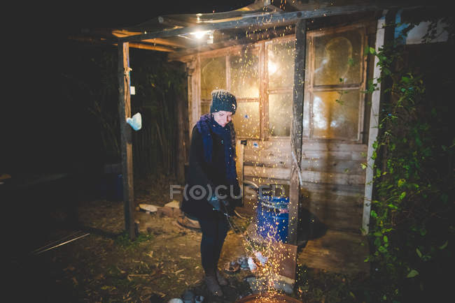 Young woman by shed waving tongs over fire causing sparks, looking down smiling — Stock Photo