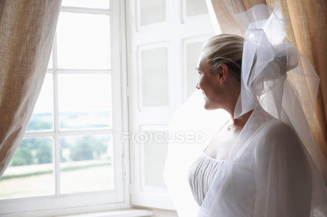 Улыбающаяся невеста стоит у окна — стоковое фото