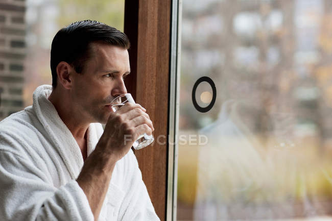 Hombre en albornoz agua potable por ventana - foto de stock