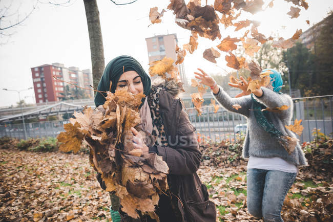 Dos amigas jóvenes juegan peleando con hojas de otoño en el parque - foto de stock
