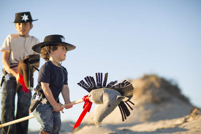 Two brothers dressed as cowboys with hobby horses in sand dunes — Stock Photo