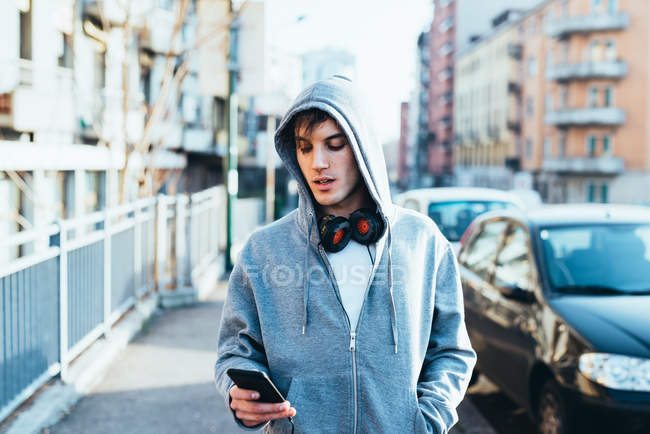 Uomo in zona urbana con cappuccio e cuffie che guardano verso il basso lo smartphone — Foto stock