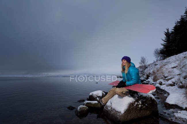 Snowboarder holding board and sitting by lake — Stock Photo