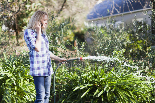 Woman on cell phone watering plants — Stock Photo
