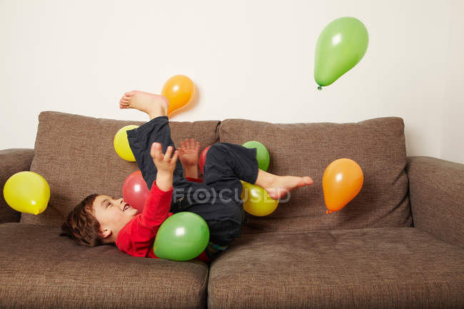 Young boy lying on sofa kicking balloons — Stock Photo