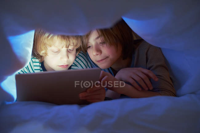 Irmãos sob edredão usando tablet digital — Fotografia de Stock