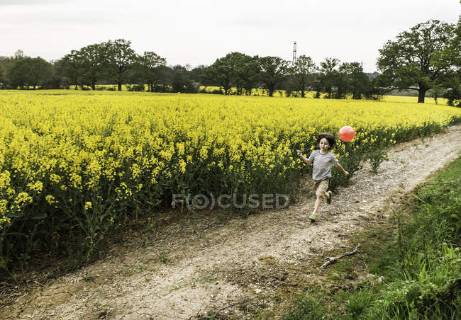 Ragazzo che corre lungo pista campo giallo fiore tirando palloncino rosso — Foto stock
