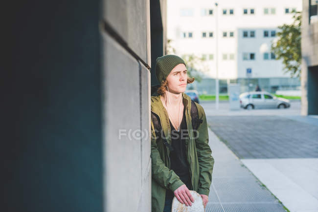 Молодий чоловік міський скейтбордист, що спирається на стіну, слухаючи музику навушників — стокове фото