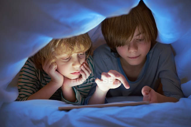 Dos chicos debajo del edredón usando tableta digital - foto de stock