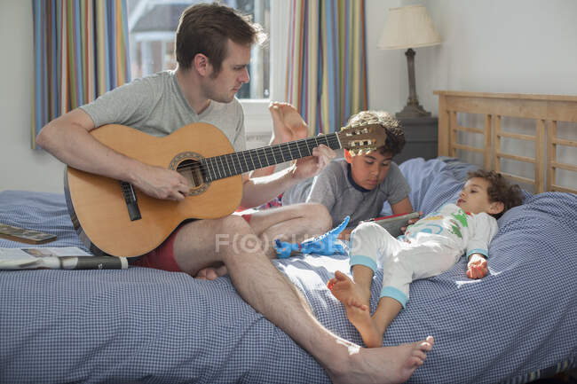 Pai tocando guitarra enquanto dois filhos relaxam na cama — Fotografia de Stock