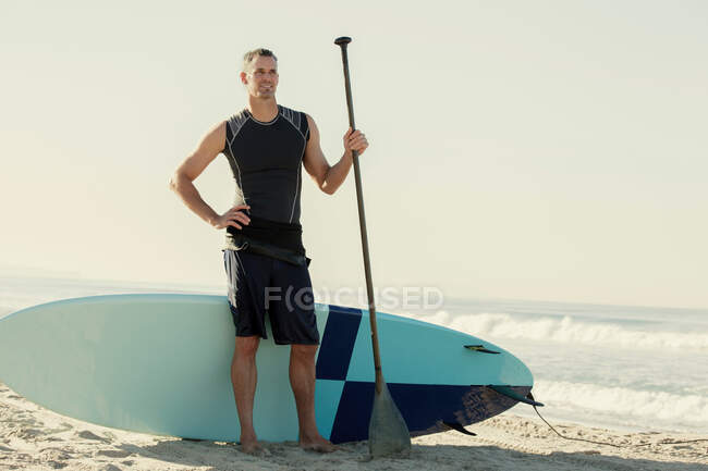 Homem adulto médio surfista de pé com prancha e remo na praia — Fotografia de Stock
