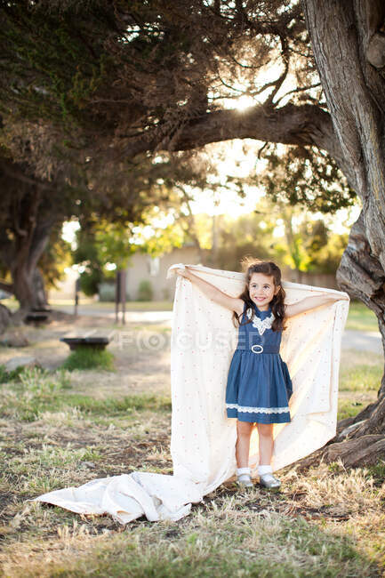 Portrait of girl holding sheet with arms out under tree — Stock Photo