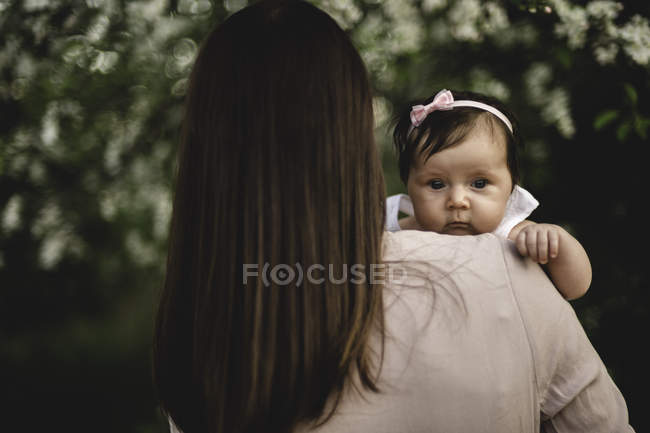 Über-Schulter-Porträt des kleinen Mädchens in den Armen der Mutter im Garten — Stockfoto
