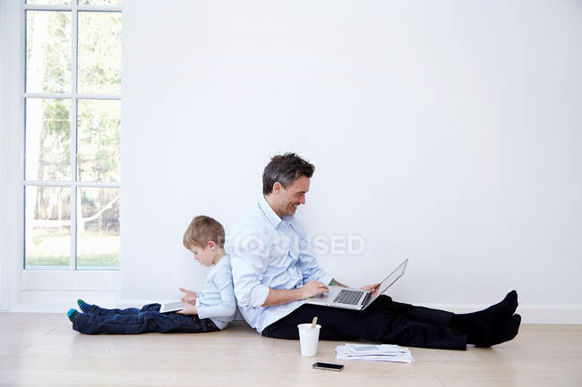 Padre e hijo sentados espalda con espalda y usando portátil y tableta digital - foto de stock