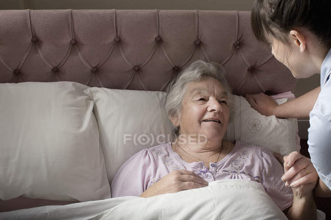 Asistente de cuidado personal charlando con una mujer mayor en la cama - foto de stock