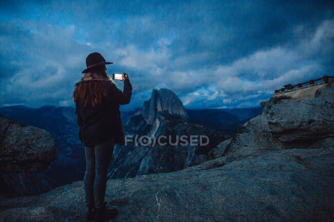 Молодая женщина фотографирует на скале с видом на Национальный парк Йосемити в сумерках, Калифорния, США — стоковое фото