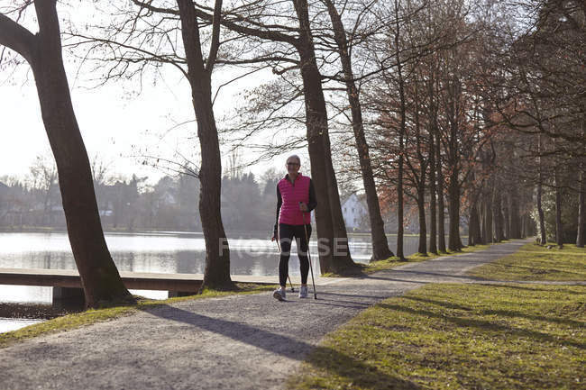 Woman nordic walking on tree lined path by pond — Stock Photo