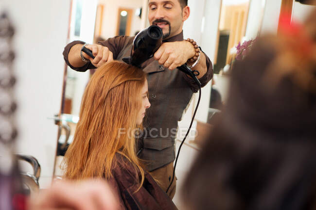Male hairdresser blow drying customer's red hair in hair salon — Stock Photo