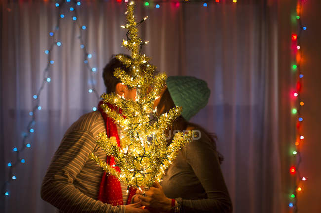 Pareja joven besándose detrás del árbol de navidad iluminado - foto de stock