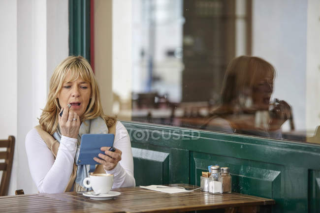 Mature woman using smartphone to apply lipstick at sidewalk cafe table — Stock Photo