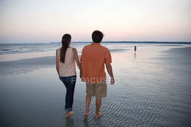 Pareja caminando en la playa con su hija en la distancia - foto de stock
