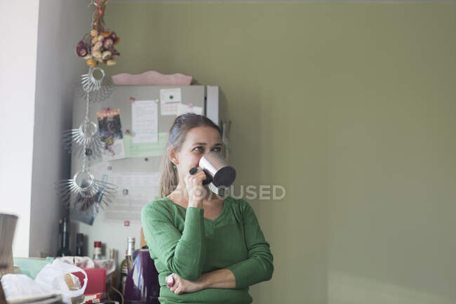 Середня доросла жінка на кухні п'є з кубку, дивлячись подалі — стокове фото