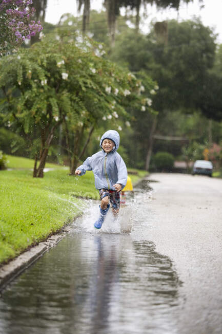 Мальчик в резиновых сапогах бежит и брызгает в лужу дождя — стоковое фото