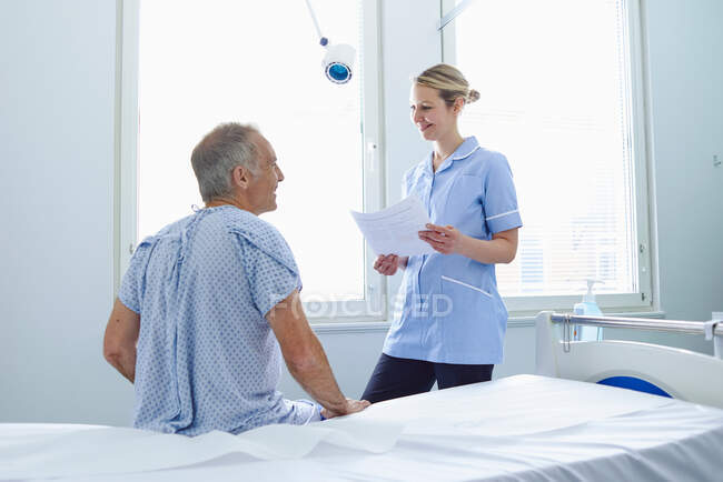 Enfermeira de pé conversando com o paciente sentado na cama do hospital — Fotografia de Stock