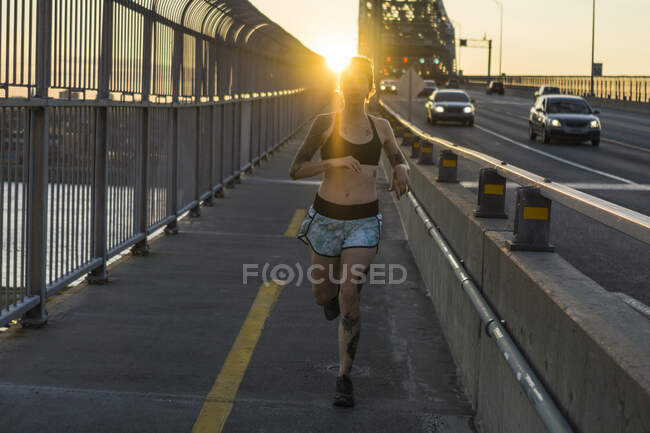 Giovane donna tatuata che corre sul ponte con il tramonto dietro — Foto stock