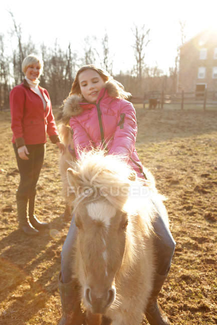 Jeune fille chevauchant poney, mère regardant par derrière — Photo de stock