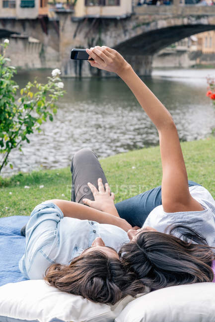 Пару лесбіянок лежачи за допомогою смартфона заволодіння selfie, навпроти мосту Понте Веккіо річки Арно, Флоренції, Тоскана, Італія — стокове фото