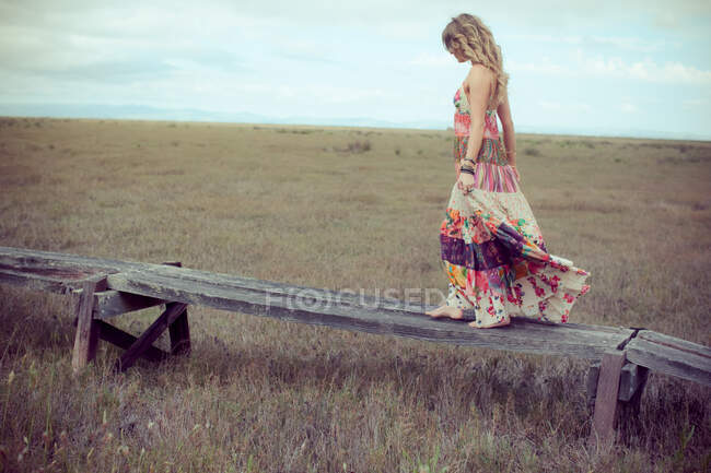 Mujer joven vestida de boho maxi caminando por el sendero elevado de madera en el paisaje. - foto de stock