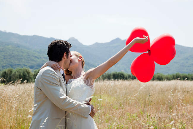 Newlyweds kissing in field with red heart shape balloons — Stock Photo