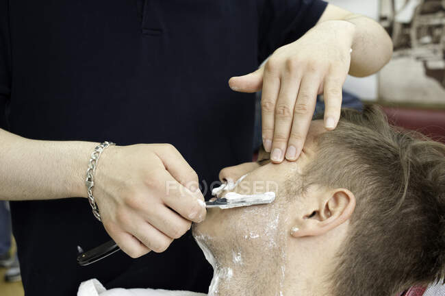 Обрезанный вид молодого человека в парикмахерской бритья клиента с прямой бритвой — стоковое фото