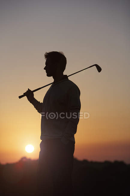 Гольфист несет клюшку для гольфа через плечо перед закатом, отводя взгляд — стоковое фото