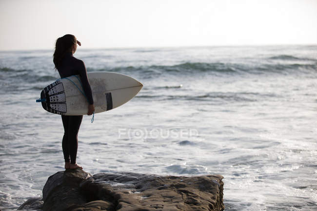 Young woman standing on rocks with surfboard — Stock Photo