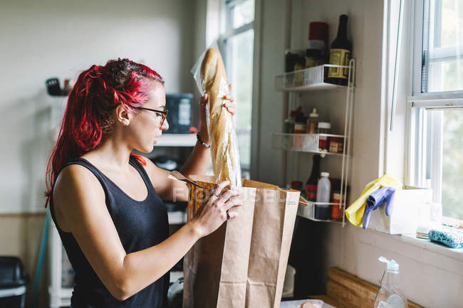 Young woman with pink hair unpacking baguette from shopping bag in kitchen — Stock Photo