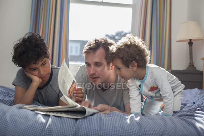 Vater und zwei Söhne relaxen im Bett, schauen auf die Zeitung — Stockfoto