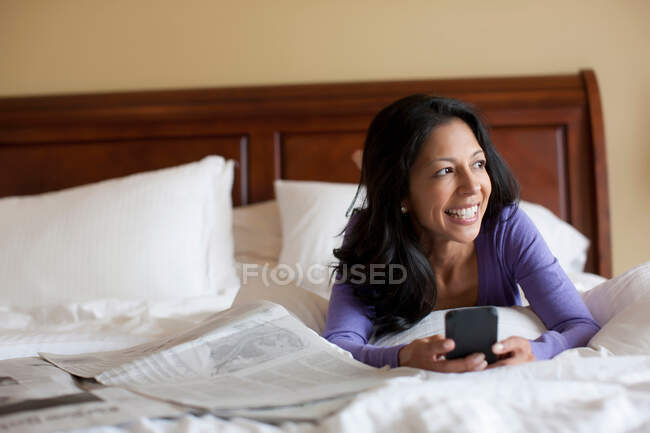 Mulher madura deitada na cama segurando telefone celular, sorrindo — Fotografia de Stock
