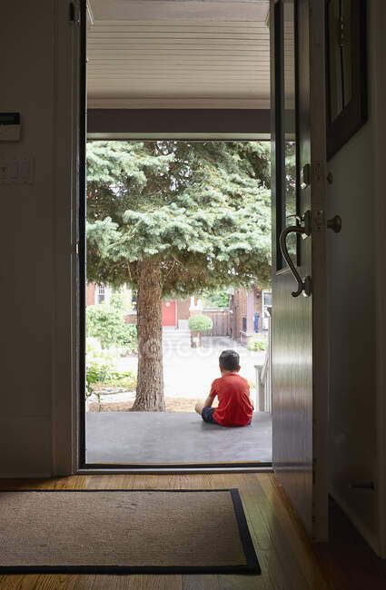 Niño sentado en el paso delantero de la casa, vista trasera - foto de stock