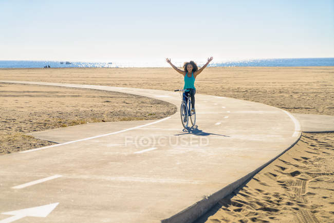 Середня доросла жінка їде на велосипеді уздовж шляху на пляжі, руки в повітрі — стокове фото