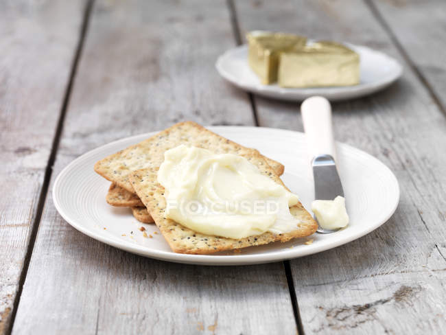 Triângulos de queijo macio untados em biscoitos quadrados com faca de manteiga na placa — Fotografia de Stock