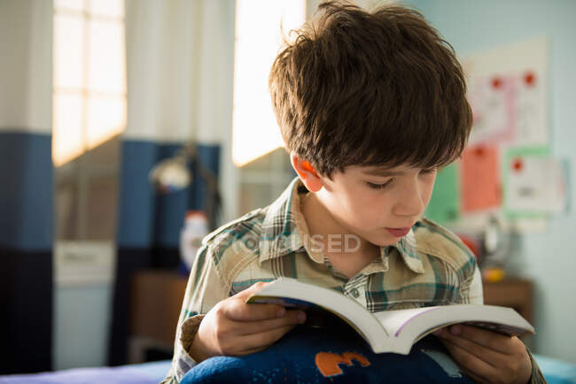 Хлопчик сидить на ліжку читає книгу — стокове фото