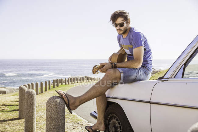 Hombre adulto sentado en el coche en la costa tocando la guitarra, Ciudad del Cabo, Sudáfrica - foto de stock