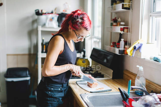 Молодая женщина с розовыми волосами режет мясо на кухне — стоковое фото