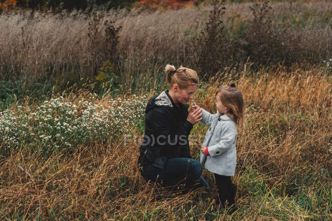 Mujer adulta mediana oliendo flores silvestres con hija pequeña en el campo de hierba larga - foto de stock