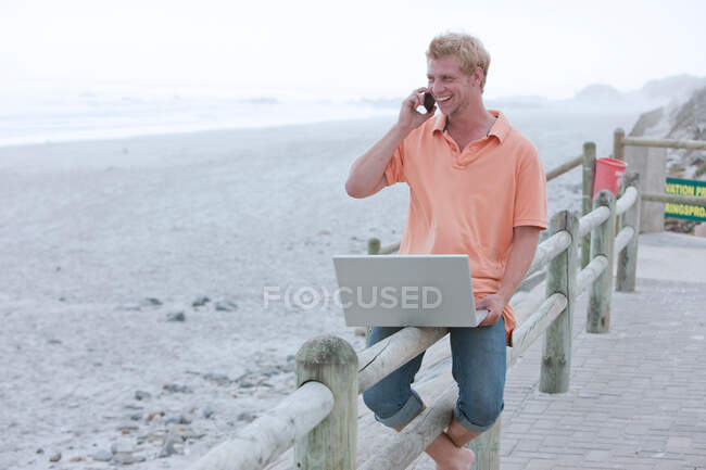 Чоловік сидить на пляжних перилах за допомогою телефону та ноутбука — стокове фото