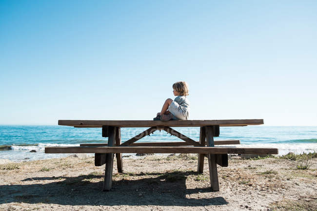 Мальчик сидит на столе для пикника на пляже — стоковое фото