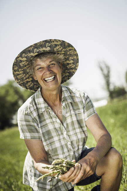 Жінка в полі в сонячному капелюсі тримає спаржу, дивлячись на камеру посміхаючись — стокове фото