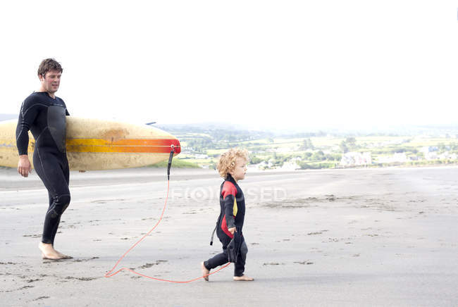 Père portant planche de surf avec fils sur la plage — Photo de stock